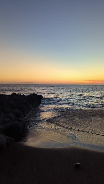 The image captures a beach scene at Pererenan, Indonesia during a sunset. The sky transitions from blue to orange, indicating the time of day. The ocean is moderately turbulent, with waves breaking on the shore. There is a rocky structure along the shoreline. The beach is a mix of wet sand where the waves are receding and darker, dry sand. A small piece of debris sits on the dry sand.
