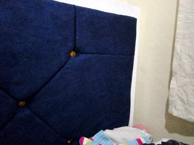 The image depicts a close-up of a tufted headboard, covered in a navy blue fabric. Diamond patterns are visible, formed by the tufts, which are secured with gold-colored buttons. The headboard is framed with a white material along the edges. In the foreground, there's a pile of colorful clothing. A beige wall is visible in the background, along with a partially seen white cloth.