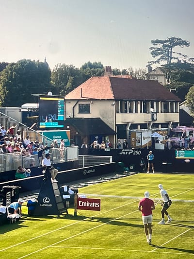 This media file captures a tennis match taking place on a sunlit grass court, likely in Brighton, United Kingdom, given the context. The image shows two male tennis players in action. One player, wearing a white cap and shirt with dark shorts, is in the process of hitting a tennis ball. The other player, dressed in a red shirt and white shorts, is positioned on the other side of the net, observing the play.
The scene is set at a tennis venue with spectators seated in tiered stands in the background. The court is meticulously maintained, with distinct white lines marking its boundaries. A net divides the court, and a tennis umpire sits in a high chair on one side. Promotional banners and signage are visible around the court, including "Emirates" and "Lexus," suggesting a professional tournament. The time of day appears to be afternoon, indicated by the bright sunlight and the long shadows cast on the grass. The weather seems pleasant and sunny, conducive to an outdoor sporting event. The overall atmosphere appears to be one of focused competition, with the players engaged in the intensity of the match. The presence of lush green trees and buildings in the background further establishes the outdoor setting.