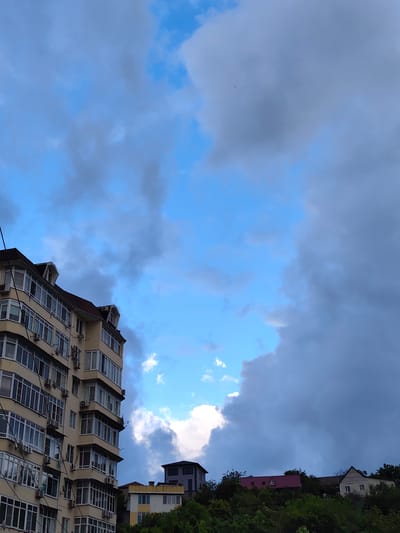 The image depicts a view from a window or balcony in the city of Oryol-Izumrud, Russia. In the foreground, a multi-story apartment building with numerous windows and air conditioning units is visible on the left side. The building is painted a light yellow color and has a reddish-brown roof. In the background, several other buildings and a lush green hillside are present. The sky is a mix of bright blue and dark, heavy clouds, suggesting an impending change in weather.