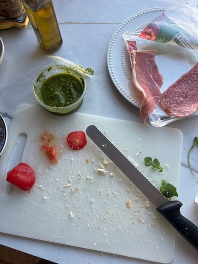 The image shows a kitchen counter with various food preparation items. A white cutting board with chopped tomatoes, breadcrumbs, and a serrated knife is in the foreground. To the left, a clear container of green pesto and a bottle of olive oil are visible. To the right, a white plate holds slices of prosciutto and salami. The scene appears to be a domestic setting, possibly in a kitchen in Brighton, United Kingdom. The lighting suggests it is daytime, and the overall impression is one of casual food preparation.