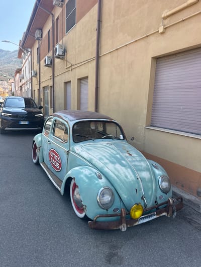A vintage, light blue Volkswagen Beetle, showing significant rust and wear, is parked on an asphalt street next to a tan building with a dark brown trim. The Beetle has white-walled tires with red rims and a circular red and white sticker on its side that reads "OTTICA MACIS." A newer black SUV with its license plate visible is parked behind the Beetle. The building has multiple windows and air conditioning units mounted on its exterior. In the background, a hilly landscape can be seen under a clear sky. The scene appears to be in a Mediterranean town, likely Muravera, Italy, given the context.