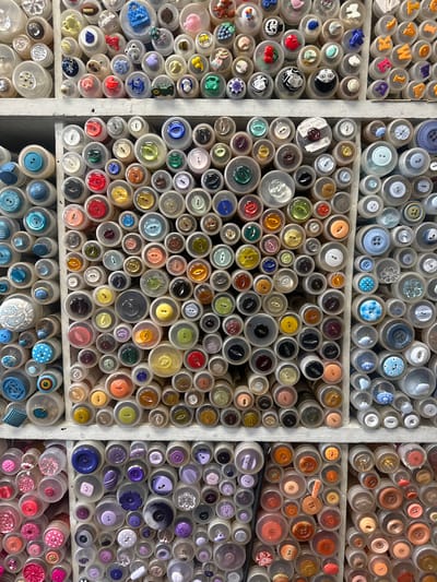 The image shows a close-up view of a collection of buttons neatly arranged in small, clear containers, organized within a set of cubbies or shelves. The buttons vary widely in color, size, and design, ranging from simple, plain buttons to more decorative ones with patterns, shapes, and embellishments. The containers are predominantly circular. The grid-like structure of the shelves and the systematic arrangement of the containers suggest an organized collection or display.
The OCR output includes text fragments such as "K", "M", "Well", "lly Hall" and "Sevenoaks" which, along with other glyphs, seem to be extracted from labels or writing possibly associated with the buttons or their containers.
Based on this image being located in Sevenoaks, United Kingdom, the collection is likely located in a crafts store or a personal collection in a home within this city.