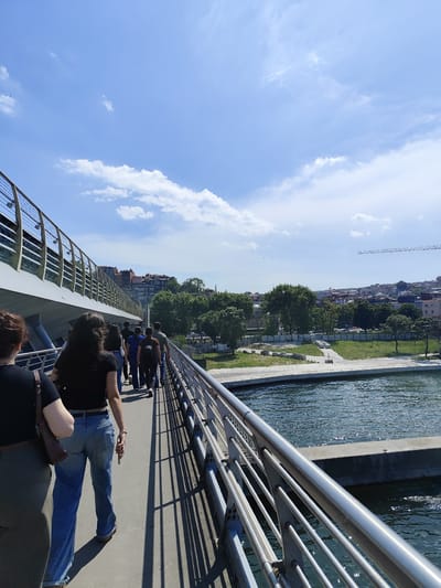 The image depicts a group of people walking across a modern bridge in Istanbul, Türkiye, likely the Golden Horn Metro Bridge. The bridge, characterized by its metallic railings and supports, spans a body of water, presumably the Golden Horn. A concrete structure is visible in the water adjacent to the bridge. In the background, the cityscape includes trees, buildings, and what appears to be a construction crane. The sky is blue with scattered clouds. The people are walking in the same direction, away from the camera, and are dressed in casual clothing. The lighting suggests it is daytime.