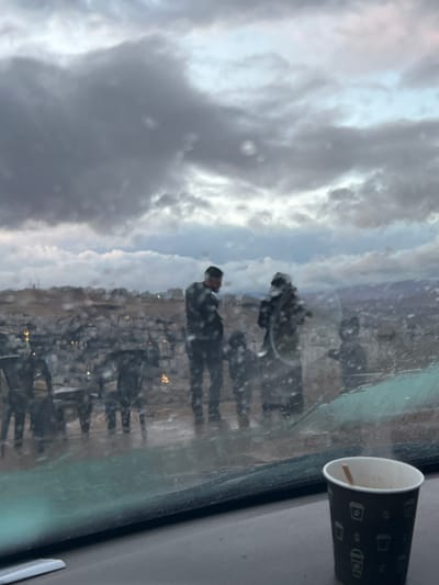 The image captures a scene in Dummar Municipality, Syria, viewed through a wet vehicle windshield. In the foreground, a dark-patterned disposable cup with a stirring stick sits on the vehicle's dashboard, to the lower right.

Outside the vehicle, the midground features four individuals on what appears to be a wet, outdoor paved area. An adult male, with a beard and dark hair, wearing a dark top, stands facing left. To his right stands another adult, clad in dark clothing and a head covering, appearing to interact with or hold a dark, cylindrical object, possibly a hookah pipe. Two smaller figures, consistent with children, are positioned between the two adults. To the left of this group, several dark plastic chairs and a table are visible on the wet ground, suggesting an outdoor seating arrangement.

The background reveals a densely built urban landscape, characterized by numerous buildings sprawling across a hillside or valley. Distant hills or mountains are discernible beyond the cityscape. The sky is heavily overcast with dark grey clouds, indicating gloomy weather conditions. The overall lighting is dim, consistent with an overcast day or twilight.