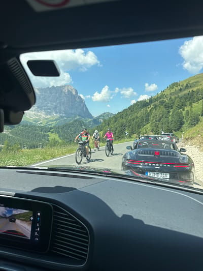 The image, taken from the interior of a vehicle, captures a scenic mountain road in Sëlva - Wolkenstein - Selva di Val Gardena, Italy. The road is populated with several cyclists and cars. In the foreground, a black convertible Porsche with a license plate reading "K-HD 318" is visible. Behind it, other cars are seen. Several cyclists are riding on the road. One cyclist, wearing a bright pink top and helmet, is on a black bicycle. Another cyclist, in a black and colorful jersey and helmet, is on a bicycle. A third cyclist, wearing a white helmet, is on a red bicycle. The road winds through a lush green mountainous landscape with trees and white wildflowers. In the background, a towering, rugged mountain dominates the skyline under a blue sky with scattered white clouds. The interior of the vehicle shows a dashboard with a screen displaying a rearview camera feed.