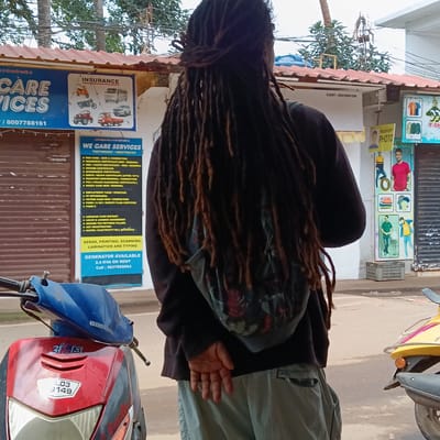 A man with long dreadlocks stands with his back to the camera in Oxel, India. He wears a black long-sleeved shirt, khaki pants, and a backpack with a floral pattern. He faces a storefront offering services, including assistance with PAN cards, residence certificates, OBC/caste certificates, and Aadhaar card reprints. Next to the storefront is a passport photo shop. Two scooters are parked on the street, one red and blue with the license plate "A.03 3149" and the other yellow. The environment is urban with overhead wires and trees.