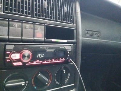 The image shows the interior of a car, likely an older model Audi due to the "Audi EC" badge visible on the dashboard. The focus is on the car's stereo system, a Pioneer brand unit with a prominent volume knob and display. The display shows "AUX" indicating it's set to an auxiliary input, and a 3.5mm audio cable is plugged into the front of the stereo. Various buttons and controls for the stereo and car functions, such as climate vents and hazard lights, are visible. The overall aesthetic suggests a slightly dated car interior, likely photographed during the day given the ambient lighting. The scene appears to be a personal, everyday moment rather than a specific event.