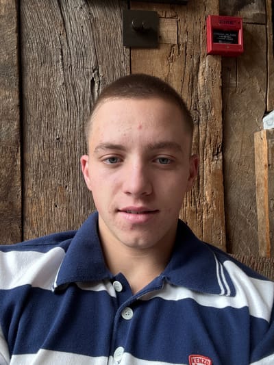 A young Caucasian male, approximately 18-25 years old, with short brown buzz-cut hair and blue eyes, is depicted from the chest up, centered in the frame. He has a fair complexion with minor dermal irregularities on his forehead and cheeks. He is wearing a navy blue and white horizontally striped polo shirt with a collar and a three-button placket featuring white buttons. A red patch with "KENZO" in white lettering is visible on the lower right portion of his shirt. He is looking directly at the camera with a neutral expression, his mouth slightly open. The background consists of a textured, aged wooden plank wall. Mounted on the upper left portion of the wall is a dark, square-shaped control panel with two circular knobs. To the upper right, a red fire alarm pull station is affixed, labeled "FIRE" and "BREAK GLASS PRESS HERE." The scene is located in Kilmersdon, United Kingdom.