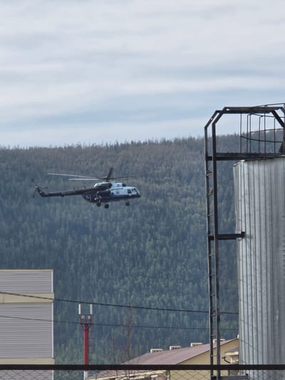 A helicopter is airborne near Lensk, Russia, with a dense forest visible in the background. Utility poles and building structures are in the foreground. The sky appears partly cloudy. The helicopter bears markings "APOGA" and "RA-24173".