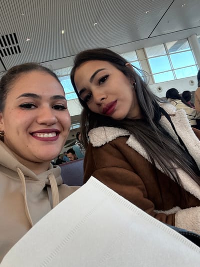 Two adult females are depicted in a close-up photograph within a bright, modern indoor environment. This location is consistent with an airport terminal, specifically Mohammed V International Airport in Nouaceur, Morocco, characterized by a high, slatted ceiling with integrated lighting and large windows revealing a bright sky. The woman on the left, foregrounded, has dark hair, dark red lipstick, and is smiling directly at the camera, displaying teeth. She wears a beige hooded sweatshirt and a visible gold hoop earring on her left ear. The woman on the right, positioned slightly behind and to her right, has long dark hair, dark red lipstick, and looks towards the camera. She wears a brown jacket with a cream-colored sherpa lining and a visible gold hoop earring on her right ear. A white, textured paper object partially obscures the lower right foreground. In the mid-background, multiple indistinct individuals are seated in rows of blue chairs.