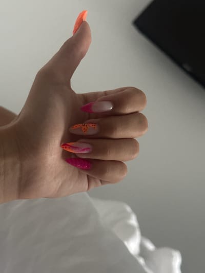 The image shows a close-up of a person's left hand, displaying almond-shaped acrylic nails. The nails are adorned with a gradient of colors, transitioning from neon orange at the tips to hot pink at the base, with some nails featuring a French tip design. One nail has a delicate floral design in orange. The hand is positioned with the thumb up, as if giving a thumbs-up. The background is out of focus, appearing to be a white or light-colored surface, possibly bedding, and a dark, blurry object in the upper right corner. The overall setting is indoor and appears casual. The image is not from Stațiunea Zoologică Marină Agigea, Romania.