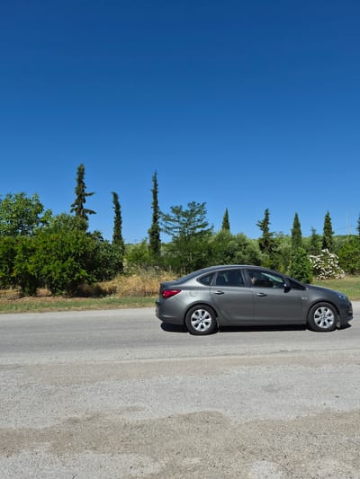 A grey sedan is traveling on a paved road under a clear blue sky. The car is captured in profile, moving from left to right. The road is bordered by dry grass and a variety of trees, including tall, slender cypress trees and bushy green foliage. The background features more greenery and a few white flowers on a bush. The setting appears to be a rural or semi-rural area in Greece, possibly near Μπομποτέϊκα, judging by the vegetation and lighting. A person's silhouette is visible through the car's window, suggesting a driver. The image is taken from a slightly elevated perspective, looking across the road.