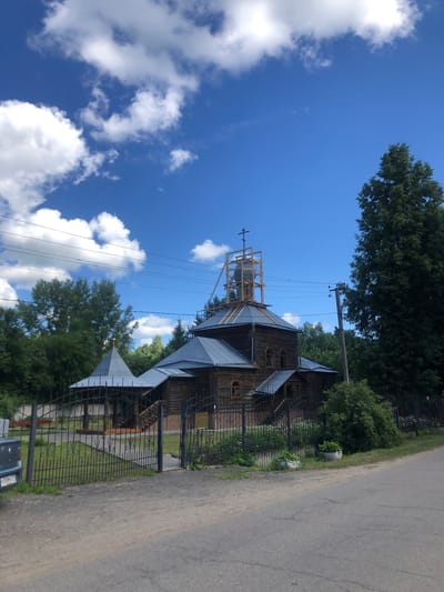 This is a photograph of the wooden Church of the Transfiguration in Izdeshkovo, Russia, on a sunny day with a bright blue sky and scattered white clouds. The church, constructed from logs, features a gray metal roof and a cross atop its belfry, which is currently under construction and surrounded by scaffolding. A smaller, octagonal structure with a metal roof is visible to the left of the main church building. The church is enclosed by a black wrought-iron fence. A paved road runs in front of the church, with a dirt path leading to the entrance. Dense green trees surround the church property, and a large, dark green coniferous tree stands to the right of the building. A power pole with wires runs across the upper portion of the image. No people are visible in the photograph.