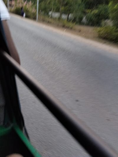 The image depicts a scene captured from a moving vehicle on a paved road in Gombato Bongwe ward, Kenya. In the immediate foreground, a dark, thick diagonal bar, likely a structural element of the vehicle, extends from the lower left to the upper right. To the left of this bar, a partial view of a person's upper arm, clad in a white sleeve, is visible, along with a prominent green strap or fabric element. The road surface, appearing grey, exhibits motion blur, indicating forward movement. In the mid-distance on the left side of the road, a solitary, blurry figure wearing dark attire is observable, seemingly walking away from the camera. The background consists of indistinct green vegetation and trees lining the road, with a light-colored pole also discernible amidst the foliage.