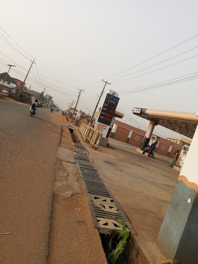 The image captures a slightly angled view of a street scene, dominated by a gas station on the right and a road with a drainage ditch on the left. The perspective is looking down the street, slightly tilted upwards.
On the left side of the frame, a paved road, appearing reddish-brown, stretches into the distance. Alongside the road, a long concrete drainage ditch is visible, covered by metal grates. In the immediate foreground, within the ditch, some green leafy vegetation (possibly corn sprouts) is growing. Utility poles with numerous overhead power lines line the road, receding into the background. A person on a motorcycle is riding away from the camera in the mid-ground on the road, and another motorcycle can be seen further down the street.
The right side of the image features a gas station. A tall, prominent digital price sign displays fuel prices. The brand name "HEKOS OIL & GAS" is visible at the top. Below it, the prices are listed:
* PMS: 828
* AGO: 790
* DPK: XXX (partially obscured, but the last digit looks like 0)
* LUSSI: 1100
Below these prices, smaller text includes "LUCKY SMART POINTS" and what appears to be phone numbers.
Adjacent to the sign is a gas station canopy structure, supported by pillars, with the "HEKOS" brand logo also visible on its edge. Behind the canopy, there are low-rise, single-story buildings painted brown with white horizontal trim. Several individuals are present at the gas station; two men are sitting on a low wall or bench near the buildings, and another person is standing further to the right.
The sky is a pale, hazy off-white or light blue, suggesting either a clear but somewhat dusty or humid day, or perhaps early morning/late afternoon with a muted light. The overall lighting is bright and even, with soft shadows. The general setting appears to be a typical urban or semi-urban environment.