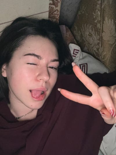 A young woman with dark hair and light skin lies down, sticking out her tongue and closing one eye. She wears a dark red shirt and a silver necklace. Her nails are long and decorated with red stripes. A hand flashes a peace sign above her face. The background includes a patterned couch, a pillow with a geometric design, and a hint of another surface.