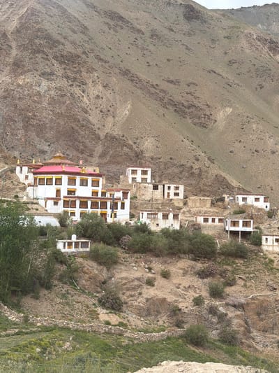 The image depicts a monastery nestled in a mountainous landscape. The monastery complex consists of several whitewashed buildings with red roofs, accented with yellow trim around the windows and cornices. Some buildings feature tiered roofs and decorative golden finials. The monastery is situated on a steep, arid hillside, with sparse vegetation consisting of low-lying shrubs and some trees. The surrounding terrain is characterized by rocky, eroded slopes and a dusty, brown-green hue. The sky is overcast. The image appears to be taken from a lower vantage point, looking up at the monastery. There are no people visible in the image. The scene suggests a remote, possibly religious, settlement in a harsh environment. The location is identified as Reru, India, which is a village in the Lahul and Spiti district of Himachal Pradesh, known for its Buddhist monasteries and rugged mountain scenery.