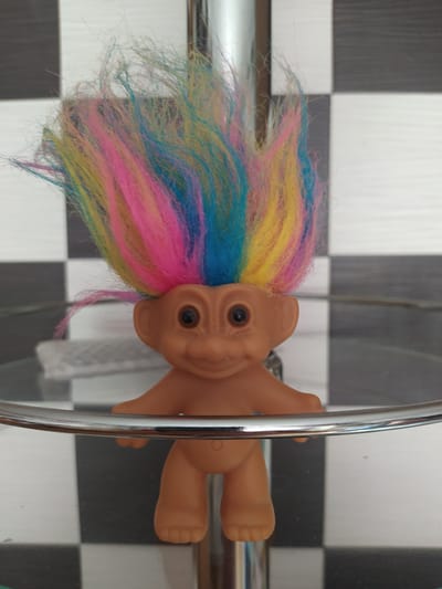 A close-up, frontal, eye-level shot features a vintage troll doll with bright, multicolored hair in shades of pink, yellow, and blue. The doll is made of tan plastic, with large round eyes, a small nose, and a subtle smile. It is standing upright. The background consists of a checkered pattern of black and white squares, likely wallpaper or tile, and a metallic support structure is visible in the foreground, partially obscuring the doll's legs and midsection. The image lacks any discernible connection to the city of Gusev, Russia, or any urban environment.