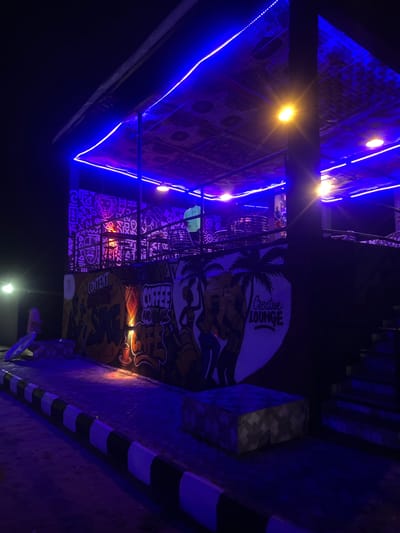 The image depicts an outdoor space at night, likely a lounge or cafe, illuminated by vibrant blue and yellow lights. The setting features a canopy or roof structure adorned with patterned fabric and string lights. A prominent mural with a tropical theme, including palm trees and stylized figures, covers a wall. Text visible in the mural reads "Creative Lounge" and "COFFEE & MORE". Other decorative elements include patterned surfaces and what appear to be tiered seating or structures in the background. The ground is paved, and a striped border runs along the edge of the paved area. The overall atmosphere is festive and inviting, suggesting a lively evening ambiance. The scene is set in Mopo-Akinlade, Nigeria.