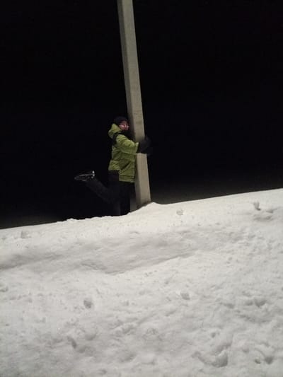 In Старый Шагирт, Russia, a nighttime outdoor scene is depicted with a person interacting with a utility pole amidst extensive snow cover. A single individual, dressed in a lime-green winter jacket, dark pants, dark beanie, and dark gloves, is embracing a light-colored, rectangular utility pole with both arms. Their left leg is bent at the knee and raised behind them. The person's face, partially visible and seemingly wearing eyeglasses, is turned towards the viewer with an apparent smile. The foreground and midground are covered in a thick layer of white snow, showing some texture and minor undulations, while the background is uniformly dark. Illumination is present on the subject and snow, contrasting with the dark background.