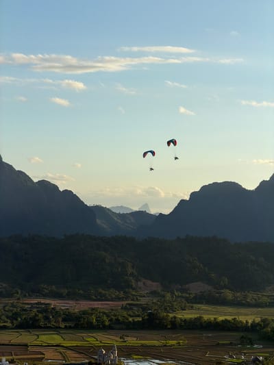 In Hong Ngern Village, Laos, three paragliders are seen in the sky over a landscape of lush green mountains and terraced fields. Two of the paragliders are closer together, with one slightly behind the other, while the third paraglider is further in the distance. The paragliders appear to be engaged in recreational activity. The environment is characterized by rolling hills covered in dense vegetation and agricultural land below. The sky is partly cloudy with a clear blue expanse. In the foreground, a cluster of buildings can be seen nestled amongst the fields, suggesting a small settlement. The overall scene depicts a tranquil rural setting with a touch of adventure in the sky.