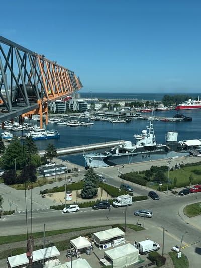 The media file shows an aerial view of a marina and waterfront area in Gdynia, Poland. In the foreground, a large, orange and gray construction crane is prominently featured, extending diagonally across the frame. Beyond the crane, the scene opens up to a vibrant harbor filled with numerous yachts and boats docked in the blue water.

Several buildings with modern architecture line the waterfront, interspersed with green spaces and trees. A road runs parallel to the harbor, with several cars visible, including a white van and a white sedan. There are also white tents or canopies set up along the road, possibly for a market or event.

In the middle ground, a large, dark gray ship with the designation "H.34." visible on its side is moored. Other vessels, including a blue and white boat, are also present in the water. The overall impression is of a bustling, well-maintained urban waterfront.

The time of day appears to be daytime, with bright sunlight casting shadows, suggesting clear weather. The clear blue sky dominates the upper left portion of the image. While no specific events are explicitly depicted, the presence of the crane suggests ongoing construction or development. There are a few people visible in the scene, mostly appearing as small figures walking or standing near the waterfront and road.

Visible text includes "H.34." on the ship and "KASA TICKETS" on a building near the waterfront. The location context provided confirms Gdynia, Poland, which aligns with the general appearance of a European coastal city. The image evokes a sense of a lively maritime hub with a mix of recreational and commercial activities.