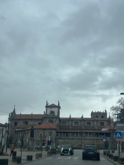 This image depicts a street scene in Lamego, Portugal, dominated by the multi-level stone structure of the Sanctuary of Nossa Senhora dos Remédios under an overcast sky.

The large building, occupying the middle ground, features a prominent central tower with a red-tiled roof and a white upper section topped by a cross. Numerous arched windows are visible across its facade, which is constructed from grey stone. Decorative finials and classical architectural elements adorn the building's various sections, all covered with red-tiled roofs. A smaller, adjacent building with a red-tiled roof and two stories stands to the left of the main structure.

In the foreground, a grey cobblestone street extends towards the background. Concrete bollards line the edges of this road. Two dark-colored passenger vehicles are present on the street, facing away from the viewer: a dark SUV in the center-left lane and a dark hatchback in the center-right lane. Several other parked cars are visible along the street to the left and right.

On the left side of the image, a lone individual, dressed in a light brown jacket, walks on a paved pedestrian area, moving away from the viewer. A dark statue on a stone pedestal is positioned in a square area to the left, in front of the smaller building. To the right, blue traffic signs are visible, including a circular sign indicating a roundabout and a rectangular sign marked "rota N2" with a pedestrian crossing symbol. Bare tree branches are partially visible on the far right. The overall environment is illuminated by diffuse light from the heavily clouded sky.