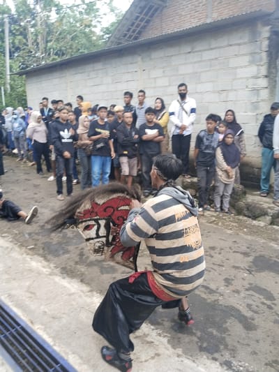 In Pendem, Indonesia, a crowd of people observes a man in a horizontally striped tan and black shirt, black pants, and red accents, squatting with a traditional horse effigy. The effigy is predominantly red, white, and black. A person lies prone on the ground to the left of the performer. A concrete block wall runs along the back of the crowd. The sky is overcast, and the street appears wet.
