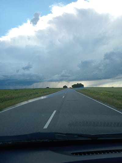 The image depicts a view from inside a vehicle driving on a paved road. The road stretches into the distance, bordered by green fields on either side. A dark, stormy cloud formation dominates the sky, with visible rain shafts extending from the clouds onto the fields in the distance. Several utility poles are scattered along the side of the road. The scene appears to be set in a rural or exurban area, possibly on the outskirts of Nikolskoye, Russia, given the presence of utility poles and open fields. The weather suggests an approaching or ongoing storm.