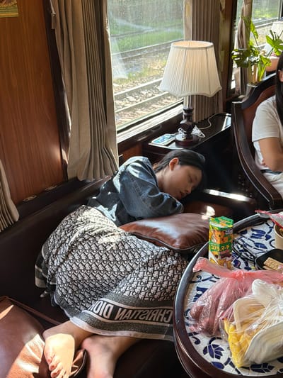 A person is sleeping on a sofa, resting their head on a brown pillow. They are covered with a patterned blanket that appears to be a scarf. A tray table with food is positioned next to the person. The setting is indoors, with a window and lamp visible in the background. Blinds are drawn on the window. Based on other details and contextual information, it may be in the city of Huế, Vietnam.