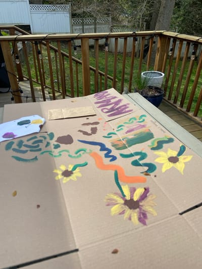 The image shows a cardboard box with a painted design on a wooden deck. The design is a simple abstract art piece with painted swirls in green, blue, orange, and purple colors. There are three painted yellow flowers on the box. There is also a brown paper bag laying on the box. The location of the image is on a deck in Rye, New York, based on the provided latitude and longitude coordinates. The wooden deck is surrounded by a wooden fence and a grassy yard. There are string lights strung up on the wooden fence. A potted plant is sitting on the deck to the right of the image. There is also a small piece of paper with a purple, green, and yellow design on it. The deck is in a residential neighborhood in Rye, New York.