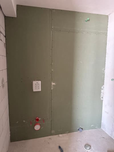In Sušćepan, Montenegro, an unfinished bathroom features walls constructed from sheets of green drywall, indicating ongoing construction or renovation. The drywall is installed in large panels, secured with visible screws, and has a rough, matte texture, suggesting it is awaiting finishing touches like joint compound and paint.
On the left drywall panel, a white recessed box with a symbol-based diagram is mounted. Below this, a red circular fixture with markings is positioned close to the floor, accompanied by two smaller red pipe-like connectors extending horizontally. Further down, a gray pipe protrudes from the wall near the floor.
The right drywall panel is largely plain, with a green pipe fixture extending from the upper section. A white pipe with a gray cap emerges from the bottom right corner of this panel. The floor is a rough concrete surface, scattered with construction debris.
Adjacent to the drywall, a wall of white concrete blocks is partially visible on the left, showing the rough texture of unfinished construction. The overall environment suggests a functional space in a state of development, with plumbing and wall structures in place but lacking aesthetic completion. The lighting is moderate, with shadows cast on the floor and walls, indicating an indoor environment with artificial or natural light sources outside the frame.