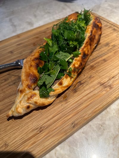 A long, boat-shaped flatbread, identified as a Pide, is positioned diagonally on a rectangular wooden cutting board. The flatbread features a golden-brown, slightly glossy crust with some darker, charred areas along its edges and surface. Its open cavity is densely packed with fresh green herbs, including prominent sprigs of parsley, dill, and mint. A brown filling, consistent with seasoned ground meat, is visible beneath the herbs. Small white crystals, likely salt, are discernible on the herb leaves. A silver-colored metal fork is partially visible, resting on the left side of the cutting board near the pointed end of the Pide. The wooden cutting board exhibits a light wood grain and multiple fine cut marks from previous use. The cutting board rests on a light-colored, mottled marble or stone countertop. The scene is illuminated by warm, directional lighting. This image depicts a food presentation in West Palm Beach, United States of America.