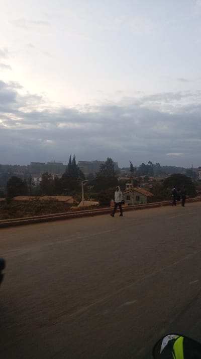 A cloudy sky hangs over a cityscape featuring buildings, trees, and a road. On the road, a person walks towards the left, while two other individuals stand on the right side. The foreground focuses on the paved road, showcasing its texture and markings. The horizon line is slightly obscured by the buildings. Overall, the scene evokes a sense of urban life under a gloomy sky.