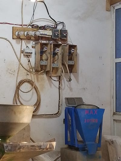 The image depicts an indoor industrial or workshop scene in Olomoda, Nigeria. A white wall serves as the background for various electrical and mechanical components.
Mounted prominently on the upper wall is a complex electrical setup. A wooden board supports three exposed incandescent light bulbs with ceramic bases, a row of multiple circuit breakers, and additional electrical switches or relays. Numerous exposed electrical wires, some bundled and others running individually, connect these components. Below the wooden board, four open, rectangular electrical enclosures are visible, each containing internal electrical contactors or relays. To the left of these, a smaller, encased electrical switch is independently mounted. A neatly coiled section of electrical cable hangs vertically below the main electrical array. Handwritten numerical markings, such as "150", are visible on the wall to the left of the coiled cable.
In the lower left quadrant, a large, light-colored metal funnel, likely part of a processing machine, is visible. Beneath its spout, a transparent tray contains granular material. In the lower right, a blue industrial hopper or feeder machine, marked with "MAX 100KG" in red lettering, sits on a concrete base. This machine incorporates a digital display unit on its upper surface. More handwritten numerical notations, including "16-1" and "16-3", are present on the white wall behind this machine. A portion of a window with a white frame and blue-tinted glass is visible along the right edge of the frame. No individuals are present in the scene, and no overt actions or interactions are occurring.