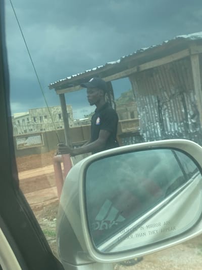 The image, taken from inside a vehicle, captures an outdoor scene in Gosa, Nigeria, rotated 90 degrees counter-clockwise. The primary subject is a dark-skinned male, wearing a dark cap and a dark t-shirt with a circular badge or logo on the left chest. He is positioned in the midground, leaning forward with his hands near a red vertical pole or pipe, seemingly engaged in an activity like working or observing.
In the foreground, the car's passenger-side mirror is prominent on the right, reflecting a blurred sky and a logo resembling the Adidas three stripes. The mirror's common safety warning "OBJECTS IN MIRROR ARE CLOSER THAN THEY APPEAR" is clearly visible in uppercase text.
Through the car window and beyond the person, a simple structure with a wooden frame and a corrugated metal roof is visible. Further in the background, several multi-story buildings, appearing to be made of concrete and possibly under construction, stand against a light blue or hazy sky. A dusty, reddish-brown dirt ground or path is seen below these structures. A thin rope or cable stretches horizontally across the lower-mid section of the frame. The scene takes place during daylight hours, under what appears to be an overcast or slightly hazy sky. The overall setting suggests a semi-urban or developing area.