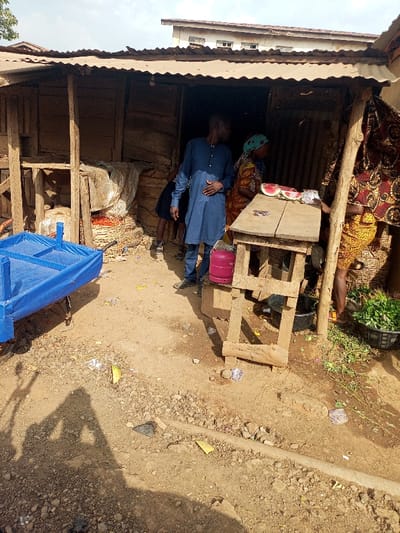 The scene depicts an outdoor market stall in Aramoko, Nigeria, under natural daylight. A rough wooden structure with a corrugated metal roof forms the stall, exhibiting walls constructed from unpainted planks and possibly corrugated sheets.
Centered, an adult male wearing a long-sleeved blue tunic stands, his left hand resting on his stomach. To his right, an adult female in a patterned dress and a light blue headscarf stands behind a rudimentary wooden counter, which displays two sliced watermelons. A pink gas cylinder is positioned on the ground adjacent to the counter. The lower portion of another individual, wearing a yellow patterned dress, is visible at the far right, standing near a black basin filled with green leafy vegetables. Inside the dim interior of the stall, large sacks of unspecified goods are discernible, along with a partially visible person wearing dark clothing on the left.
In the foreground, a blue metal pushcart or wheelbarrow with a fabric-lined bed rests on the dry, uneven dirt ground. The ground is littered with various debris, including plastic wrappers, paper, and organic waste, with a light green fruit rind visible near the center. Distinct shadows, including that of the photographer, are cast on the ground. A multi-story building with a light-colored facade is visible in the distant background, beneath a hazy sky.