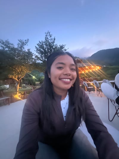 A young woman with dark hair and dimples smiles into the camera in front of a setting of outdoor dining tables and huts in Tu Le, Vietnam. The woman is wearing a dark brown blazer over a white polka-dotted shirt. The background shows a thatched-roof hut illuminated by string lights, and a mountainous landscape shrouded in dusk. Several empty white chairs are visible around tables. The overall impression is a pleasant evening at an outdoor restaurant.