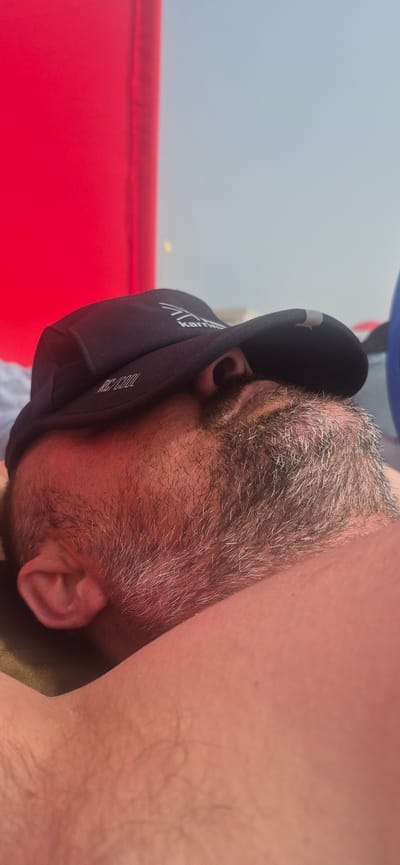 The image shows a close-up of a man wearing a black baseball cap. The cap is pulled down, covering most of his face, but a portion of his beard and ear are visible. The beard is graying and appears somewhat unkempt. The man seems to be lying down, possibly sleeping, as his eyes are not visible and his head is angled downwards. In the background, a large bright red object is visible on the left side of the image, and a pale blue sky or background is on the right. The lighting suggests it is daytime. The setting appears to be outdoors, possibly near a beach or a recreational area, given the relaxed posture of the subject and the bright, open background. Without further context, it is not possible to definitively determine the exact location within Póvoa de Varzim, Portugal.