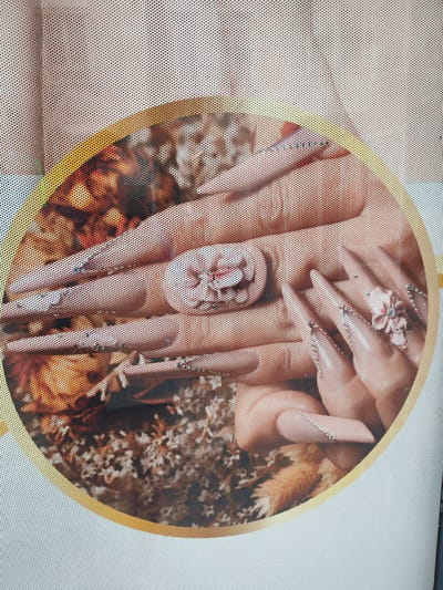 The image shows a close-up of hands with long, embellished fingernails, possibly acrylic or gel extensions, decorated with rhinestones and floral designs. The hands appear to be resting on a textured surface that includes dried flowers or foliage. The image is overlaid with a perforated pattern, suggesting it is a photograph of a window display or advertisement. The specific location in the city of Rosenheim, Germany is not discernible from the visual information provided.