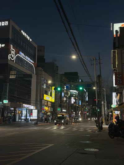 This media file captures a nighttime scene on a street in Uijeongbu-si, South Korea. The image is oriented vertically, looking down a street lined with various businesses, many of which are illuminated by neon signs.
The main subjects are the buildings with their vibrant signage, the street itself with crosswalk markings, and a few visible objects like streetlights, utility poles, and traffic signals. There are also glimpses of people, some walking along the sidewalk and others near a motorcycle, suggesting a lively urban environment. One individual is visible riding a motorcycle, adding a dynamic element to the scene.
The time of day is clearly night, with the dark sky contrasting sharply with the brightly lit signs. The weather appears to be clear, as there are no visible signs of rain or fog. The presence of Korean text on the signs and street signs definitively places the location in South Korea, specifically Uijeongbu-si as indicated by "시민로131번길" (Simin-ro 131beon-gil).
Notable details include the dense collection of signs advertising a variety of services, such as "서울한의원" (Seoul Korean Medicine Clinic), "명보치과" (Myeongbo Dental Clinic), "강원도 옥수" (Gangwondo Corn), and "노래방" (Karaoke), indicative of a commercial district. The overall mood is one of urban nightlife, with the bright lights creating a bustling and energetic atmosphere.