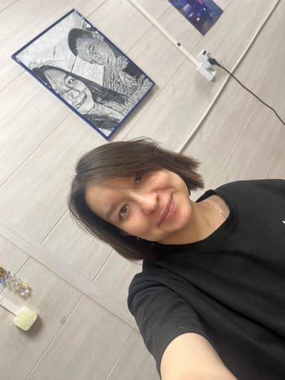 An indoor scene in Kozlovka, Russia, shows a smiling woman with short, dark hair, wearing a black long-sleeved shirt and a thin gold-colored necklace, looking directly at the camera. Her right arm is visible in the lower right. The background features light-colored wall paneling. Affixed to the upper left wall is a rectangular framed artwork with a dark blue border, depicting a greyscale image of two individuals: a smiling woman in the foreground and a man behind her. To the upper right, a white multi-outlet power strip with a black cable plugged in is mounted on the wall, with a partial view of another framed item showing dark blue and purple elements above it. In the lower left, a decorative object with clear, faceted components is partially visible, beside a small, light-colored square object.
