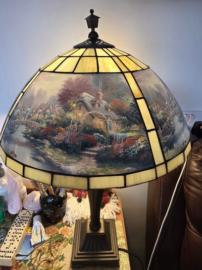 A large, illuminated Tiffany-style table lamp dominates the foreground, placed in an indoor setting in Cwmbrân, United Kingdom. The lampshade features numerous segmented panels of yellowish-orange stained glass interspersed with larger, painted glass panels. These painted sections depict idyllic, detailed landscapes, primarily featuring quaint cottages with glowing windows, lush gardens bursting with colorful flowers, arched bridges over water, and winding paths, reminiscent of a pastoral scene. The lamp's base, partially visible, is dark and appears to be made of metal or resin, with decorative fluting.
The lamp sits on a cluttered surface, likely a side table. Visible items on the table include a stack of dominoes, various bottles and tubes of toiletries or medicine (one partially showing "ДИКА" in Cyrillic, and another "and find tart"), a pair of reading glasses resting on a patterned fabric, a black glossy bird-shaped decorative object, and a white, textured, crocheted item.
In the background, a brown leather armchair or sofa is visible, with a white charging cable draped across it. A plain light-colored wall forms the upper left background. The lamp itself provides a warm, inviting glow, suggesting it is evening or that the room is dimly lit, contributing to a cozy and domestic atmosphere.