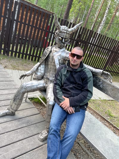 In Tal'tsy, Russia, a man in sunglasses and a black and green zip-up hoodie, blue jeans, and sneakers sits on a metal bench next to a large, detailed metal sculpture of a horned, armored figure. The sculpture is made of many small metal pieces riveted together, forming a seated warrior with a helmet, cape, and metal shoes. The man holds a black smartphone in his right hand. They are positioned on a wooden boardwalk in an outdoor setting with a wooden fence and trees visible in the background. The ground around the boardwalk is covered in small stones and grass.