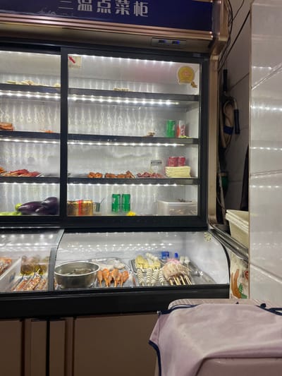 The image displays an illuminated commercial food display refrigerator located within what appears to be a small eatery or street food vendor establishment in Dali City, China. The primary subject is a tall, glass-fronted refrigeration unit featuring a blue header panel with Chinese characters that translate to "ordering display cabinet." The interior is brightly lit by LED strips along each shelf.

The upper section of the refrigerator contains several shelves. The second shelf from the top holds trays of various food skewers and individual green (Sprite) and red (Coca-Cola) soda cans. The third shelf displays additional skewers, pieces of raw meat, and stacks of flat, white food items, alongside more red soda cans. The bottom shelf of this section features two dark purple eggplants, green vegetables, a golden container, and two green soda cans, with a clear plastic container visible on the far right. A golden award or certification seal is affixed to the inside of the upper right glass panel.

Below the main display, an open-top chilled section presents numerous food items arranged on skewers for customer selection. From left to right, this section includes trays with prepared foods, a metal bowl containing grey items, skewers with orange-brown items (likely shrimp or processed meat), multiple skewers of white, cylindrical, and spherical processed food items (such as fish balls or tofu), and larger white/pinkish skewers. Small plastic water bottles are also visible among these items.

To the right of the display unit, a reflective, possibly tiled or metallic wall segment is visible. A dark strap or belt hangs vertically on this wall, positioned above stacked white plastic containers. In the immediate foreground, the edge of a counter or table draped with a light-colored cloth (possibly pinkish-purple) with a blue border is partially visible. No people are present in the scene, and no specific actions or interactions are depicted.