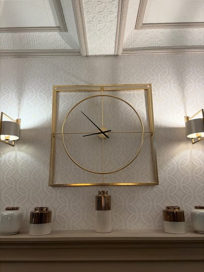 The image shows a wall in what appears to be an interior space, with decorative elements. On the left side of the image, there is a portion of a wall with raised, ornate decorative panels and molding. The central part of the image is dominated by wallpaper with a repeating pattern of interconnected, teardrop-like shapes. Mounted on this wallpaper is a square-framed clock with a gold-colored finish. The clock face is mostly open with gold-colored hands that indicate a time around 7:20. Above and below the clock are light fixtures with metallic and glass elements, while to the right of the clock are vertically aligned light fixtures with a similar aesthetic. The overall color scheme is light, with creams, whites, and gold accents. There are no people visible in the image. The location of Glenridding, United Kingdom, is not directly verifiable from the information present in the image. Therefore, no information can be provided that explicitly supports the image being located in Glenridding, United Kingdom.
