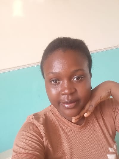A forensic examination of the image reveals a medium close-up portrait of an adult female in an indoor setting within OGBOKO, Nigeria. The individual possesses dark skin, short, dark, closely-cropped hair, and directly faces the camera with a neutral expression. She wears a light brown, ribbed, short-sleeved top. Her left hand is raised, with fingers curled beneath her chin and thumb resting along her jawline. The background comprises a two-toned wall, featuring an upper section of light beige and a lower section of turquoise, with a horizontal demarcation line visible at shoulder level. Lighting is uniform, illuminating the subject's face. No specific interior identifiers are present to determine the precise location within Ogbolo beyond it being an interior space.
