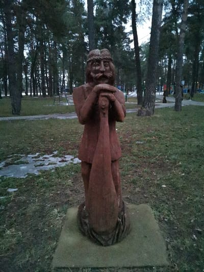 This image depicts a reddish-brown wooden statue of a male figure situated in a park in Masty, Belarus. The statue, appearing aged and carved from a single piece of wood, portrays a man with a prominent mustache and beard, and a textured head covering resembling a wreath or braided hair. His hands are clasped atop a large, elongated, tapering object, consistent with a fish or a stylized club, held vertically before the figure. The statue rests on a square, light grey concrete plinth partially embedded in the earth.
The immediate ground surrounding the statue consists of sparse, dull green grass interspersed with irregular patches of melting snow or ice, revealing dark, wet soil. A dark paved pathway is visible to the left and recedes into the background on the right. The background is dominated by a dense stand of mature conifer trees, primarily pines, with dark trunks and evergreen foliage extending upwards. Distant, indistinct playground structures, including slides and swings, are visible through the trees in the far background to the right. The scene is illuminated by diffuse, low-level light consistent with an overcast sky during a cooler season. No living individuals are present.