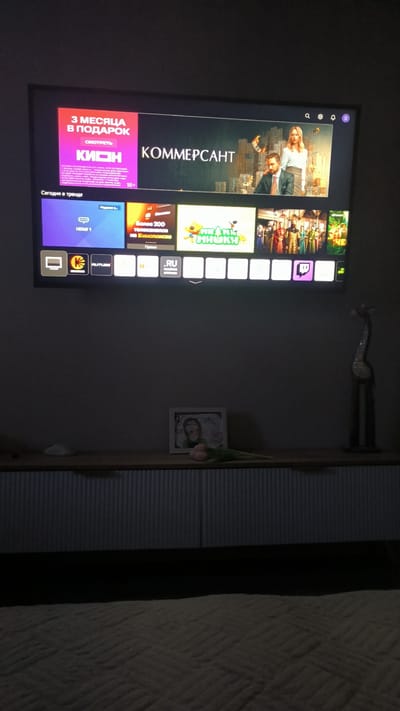 The image shows a television screen displaying various media content, including a movie or show titled "КОММЕРСАНТ" and several smaller preview tiles. The television is mounted on a wall in a dimly lit room. Below the television, a wooden cabinet with a ribbed facade is visible. On top of the cabinet, a framed photo with a handwritten message, a small white object (possibly a remote control or mouse), and a bouquet of pink tulips are placed. To the right of the cabinet, a tall, abstract sculpture resembling a giraffe is positioned. The floor in front of the cabinet is covered with a textured rug. The overall scene suggests a home entertainment setup. There is no visible indication of the specific location in Kovrov, Russia.